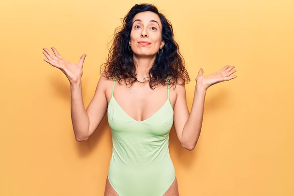 Young beautiful hispanic woman wearing swimwear clueless and confused with open arms, no idea and doubtful face.  - Photo, Image