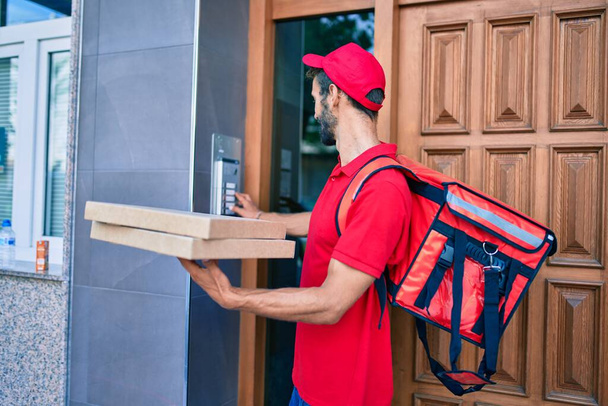 Caucásico repartidor hombre vistiendo uniforme rojo y entrega mochila sonriente feliz al aire libre sosteniendo caja de pizza - Foto, Imagen