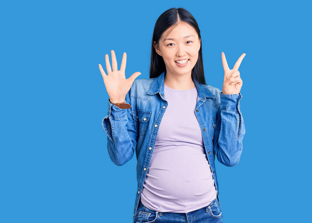 Genç, güzel Çinli bir kadın hamile. Bebek bekliyor ve yedi numaralı parmaklarıyla işaret ediyor. Kendinden emin ve mutlu bir şekilde gülümsüyor..  - Fotoğraf, Görsel