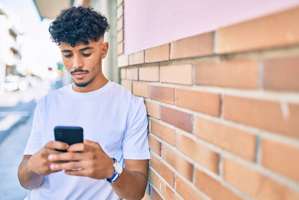 Young arab man with serious expression using smartphone leaning on the wall. - Foto, afbeelding