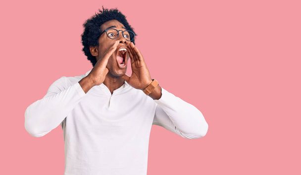Όμορφος Αφροαμερικάνος με αφρικάνικα μαλλιά φορώντας καθημερινά ρούχα και γυαλιά φωνάζοντας θυμωμένος δυνατά με τα χέρια πάνω από το στόμα  - Φωτογραφία, εικόνα