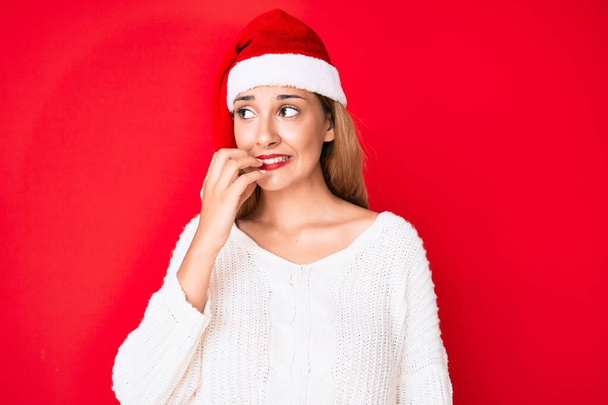 Młoda brunetka ubrana w świąteczny kapelusz wyglądająca na zestresowaną i zdenerwowaną z rękami obgryzającymi paznokcie. niepokój.  - Zdjęcie, obraz
