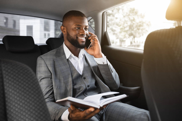 Веселый черный менеджер разговаривает по телефону, держит ноутбук в машине - Фото, изображение