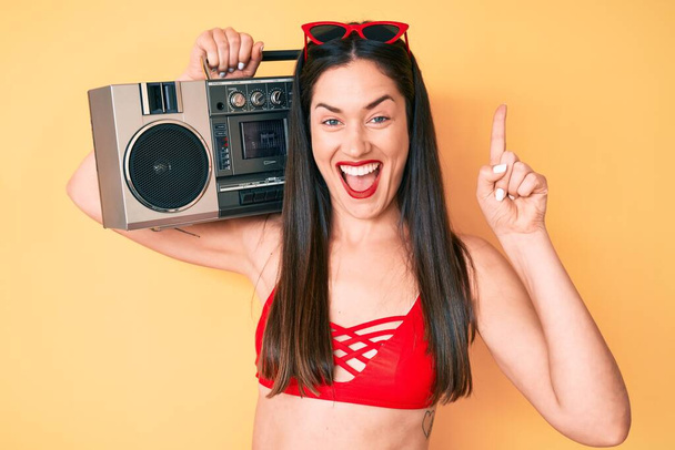 Young beautiful caucasian woman wearing bikini and holding boombox surprised with an idea or question pointing finger with happy face, number one  - Photo, Image
