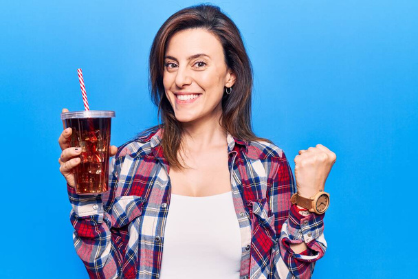 Nuori kaunis nainen juo lasillinen cola juomaa huutaa ylpeä, juhlii voittoa ja menestystä hyvin innoissaan kohotettu käsi  - Valokuva, kuva