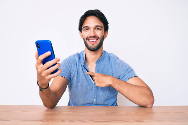 Akıllı telefon kullanan yakışıklı İspanyol adam masanın üzerinde oturmuş mutlu ve gururlu bir kendini işaret ediyor.  - Fotoğraf, Görsel