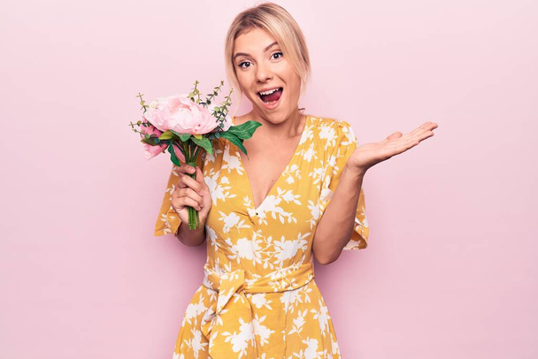 Jonge mooie blonde vrouw met boeket bloemen over geïsoleerde roze achtergrond vieren prestatie met een vrolijke glimlach en winnaar uitdrukking met opgeheven hand - Foto, afbeelding