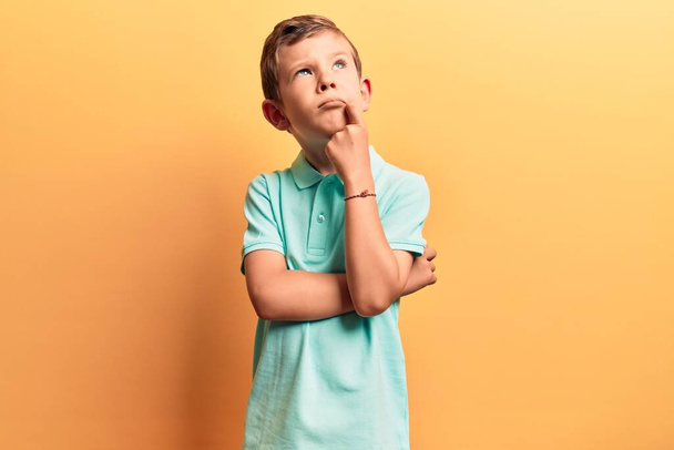 Χαριτωμένο ξανθό παιδί που φοράει καθημερινά ρούχα και σκέφτεται συγκεντρωμένα στην αμφιβολία με το δάχτυλο στο πηγούνι και αναρωτιέται  - Φωτογραφία, εικόνα