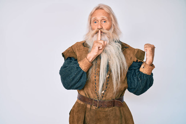 Vieil homme âgé aux cheveux gris et à la barbe longue portant un costume traditionnel viking demandant de se taire avec le doigt sur les lèvres pointant la main sur le côté. silence et concept secret.  - Photo, image