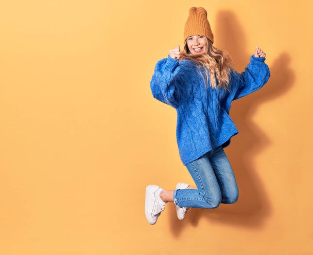 Young beautiful woman wearing casual winter clothes smiling happy. Jumping with smile on face doing winner gesture with fists up over isolated yellow background - 写真・画像