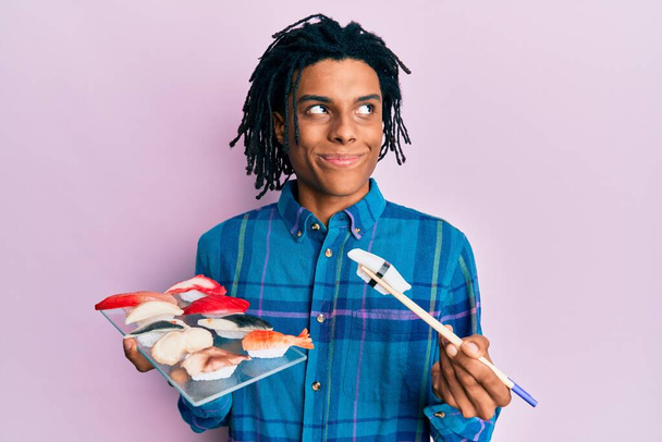 Молодий афроамериканець їсть суші, використовуючи палички, що посміхаються, дивлячись убік і дивлячись у майбутнє,.  - Фото, зображення