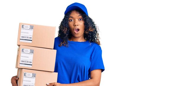 Mujer afroamericana joven sosteniendo paquete de entrega asustada y sorprendida con la boca abierta para sorpresa, cara de incredulidad  - Foto, imagen