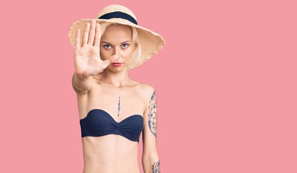 Jonge blonde vrouw met tatoeage in bikini en zomerhoed doen stoppen met zingen met palm van de hand. waarschuwingsuitdrukking met negatief en ernstig gebaar op het gezicht.  - Foto, afbeelding