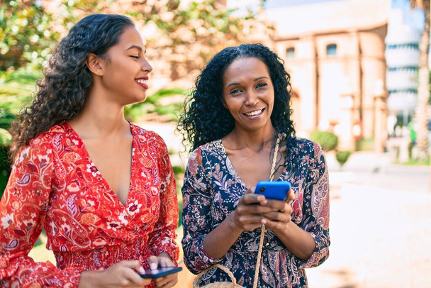 アフリカ系アメリカ人の母親と娘が公園でスマートフォンを使って笑顔で, - 写真・画像
