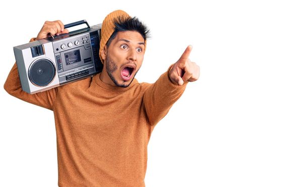 Handsome latin american young man holding boombox, listening to music pointing with finger surprised ahead, open mouth amazed expression, something on the front  - Photo, Image