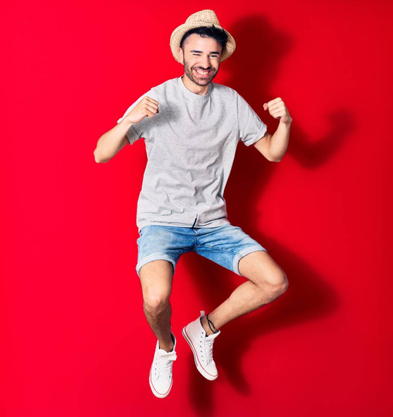 Νεαρός όμορφος Ισπανός που φοράει καθημερινά ρούχα και καλοκαιρινό καπέλο χαμογελώντας χαρούμενος. Άλμα με χαμόγελο στο πρόσωπο γιορτάζει με γροθιές πάνω από απομονωμένο κόκκινο φόντο. - Φωτογραφία, εικόνα