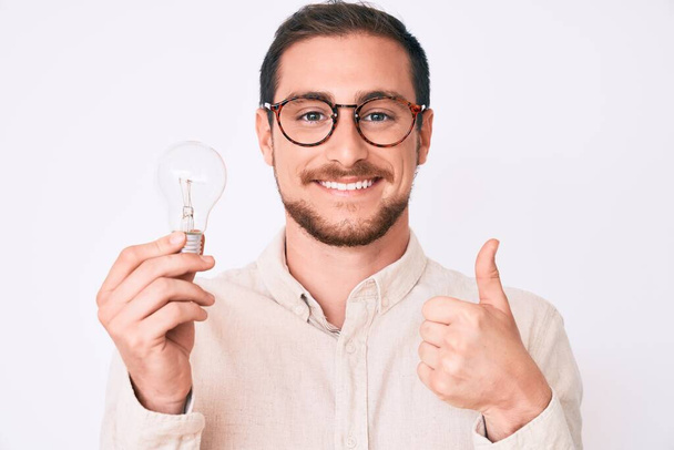 Young handsome man holding lightbulb for inspiration and idea smiling happy and positive, thumb up doing excellent and approval sign  - Foto, imagen