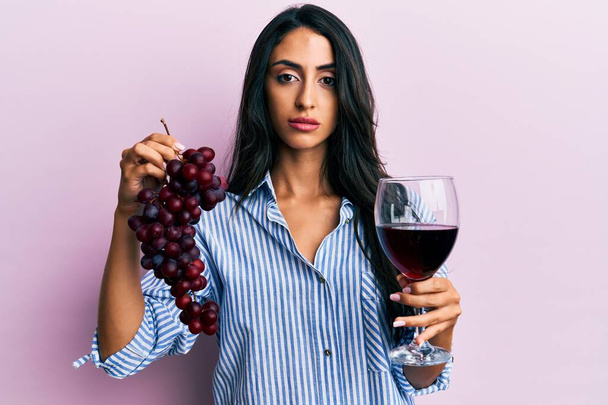 Gyönyörű spanyol nő kezében ág friss szőlő és vörösbor ellazult komoly kifejezés az arcon. egyszerű és természetes nézi a kamerát.  - Fotó, kép