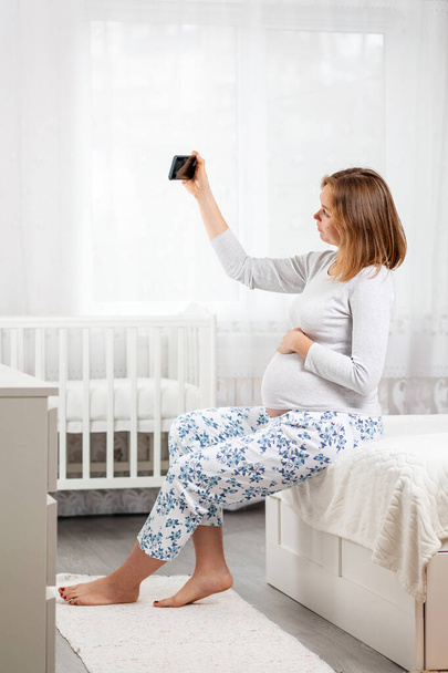 Una joven embarazada está sentada en una cama y tomando una selfie en su teléfono inteligente. Concepto de redes sociales y embarazo. Vertical y de cerca. - Foto, imagen
