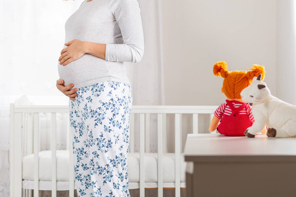 A pregnant Caucasian woman stands in the children's white room and hugs her tummy. In the background, a large window and a cradle. Close up. - Foto, immagini