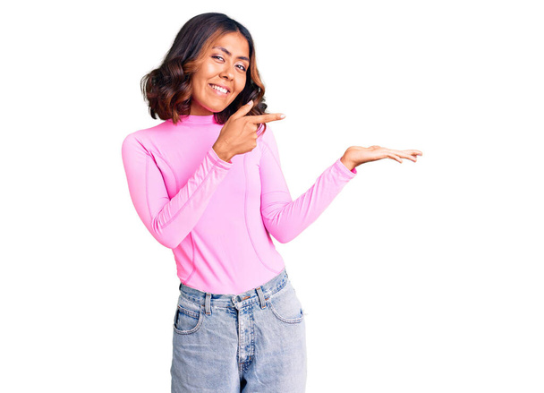 Mladý krásný smíšený závod žena na sobě růžová košile ohromen a usmívá se na kameru při prezentaci s rukou a ukazuje prstem.  - Fotografie, Obrázek