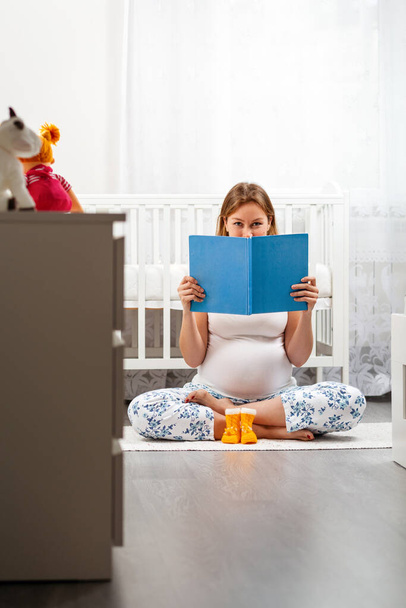 Een jonge zwangere vrouw zit op de vloer naast baby laarzen en houdt een boek bij haar gezicht. Kinderkamer op de achtergrond. Concept van zwangerschap en gelukkig moederschap. - Foto, afbeelding