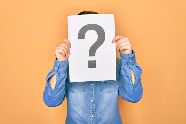 Woman covering face holding paper with question mark symbol. Standing over isolated yellow background - Photo, Image