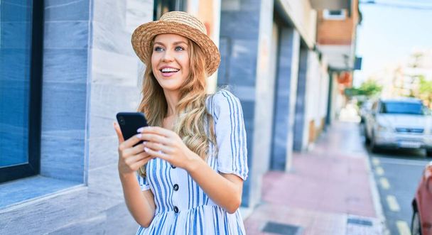 夏の帽子を身に着けて休暇中の若い美しいブロンドの女性は幸せな笑顔。街の通りでスマートフォンを使って笑顔で立って. - 写真・画像