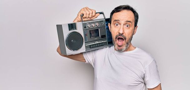 Middle age handsome man listening vintage boombox over isolated white background scared and amazed with open mouth for surprise, disbelief face - Фото, изображение