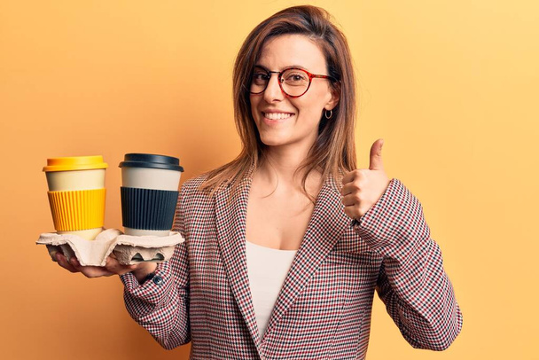 Young beautiful woman wearing business clothes holding takeaway cup of coffee smiling happy and positive, thumb up doing excellent and approval sign  - Φωτογραφία, εικόνα