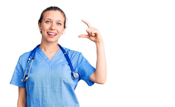 Young beautiful blonde woman wearing doctor uniform and stethoscope smiling and confident gesturing with hand doing small size sign with fingers looking and the camera. measure concept.  - Photo, Image