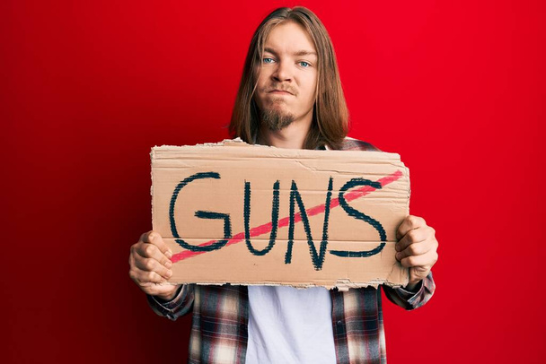 Handsome caucasian man with long hair holding no guns warning banner skeptic and nervous, frowning upset because of problem. negative person.  - Photo, Image