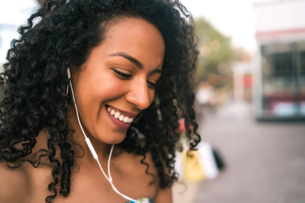 Portrait of young Afro american latin woman listening to music with earphones outdoors in the street.  - Foto, imagen