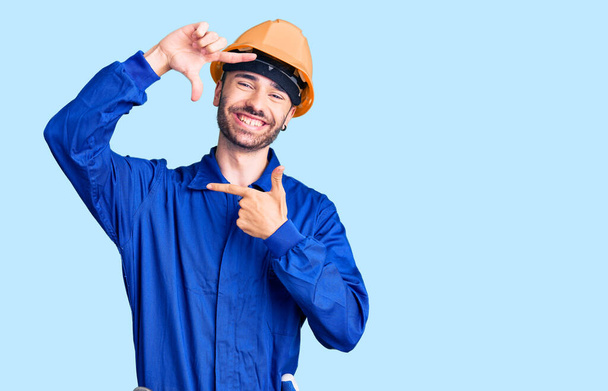 労働者の制服を着た若いヒスパニック系の男が幸せな顔をした手と指で笑顔のフレームを作ります。創造性と写真の概念.  - 写真・画像