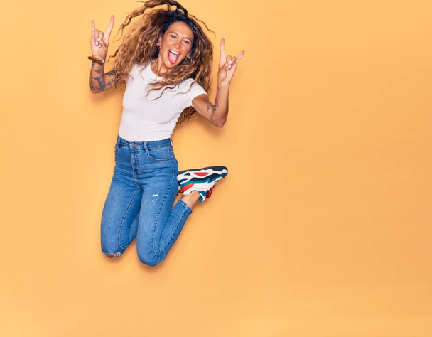 Young beautiful woman with tattoo wearing casual clothes smiling happy. Jumping with smile on face doing rocker sign over isolated yellow background - Photo, Image