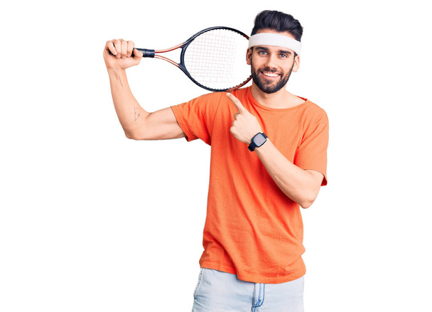 Молодий красивий чоловік з бородою грає в теніс, тримає ракетку, усміхаючись щасливо вказуючи рукою і пальцем
  - Фото, зображення