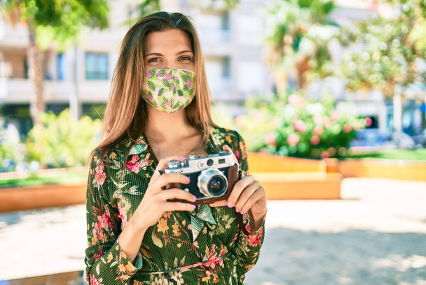Güneşli bir günde beyaz güzel bir kadın Coronavirus güvenlik maskesi takıp klasik kamera kullanarak fotoğraf çekiyor. - Fotoğraf, Görsel