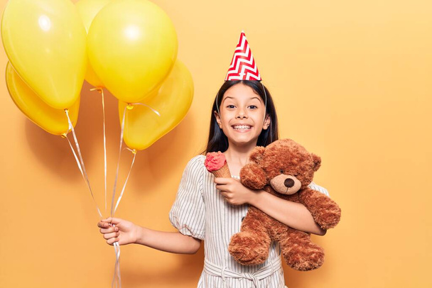 Urocza latynoska dziewczynka w urodzinowym kapeluszu uśmiechnięta. Stojąc z uśmiechem na twarzy trzymając balony i pluszowego misia jedząc lody na odizolowanym żółtym tle - Zdjęcie, obraz