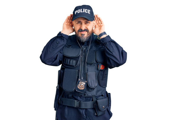 Νεαρός όμορφος άντρας με αστυνομική στολή προσπαθεί να ακούσει και τα δύο χέρια σε μια χειρονομία αυτιού, περίεργος για κουτσομπολιά. πρόβλημα ακοής, κωφός  - Φωτογραφία, εικόνα
