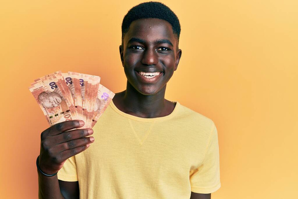 南アフリカのブランドの紙幣を持っている若いアフリカ系アメリカ人男性は、前向きな笑顔で立って笑顔で歯を見せています  - 写真・画像