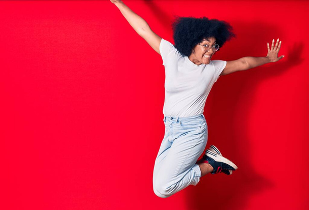 Young beautiful african american woman wearing casual clothes and glasses smiling happy. Jumping with smile on face celebrating with fists up over isolated red background - Foto, afbeelding