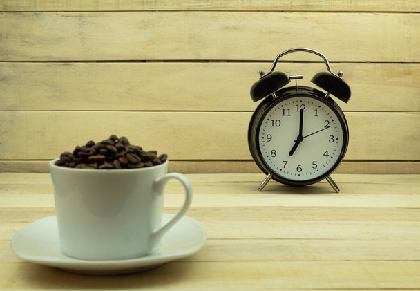 Hälytyskello ja paahdetut kahvipavut valkoisessa kupissa taustana. Tekstin malli Kello näyttää seitsemän aamulla - Valokuva, kuva
