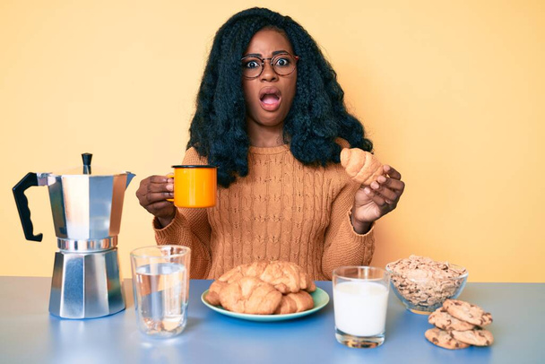 Giovane donna afroamericana che fa colazione tenendo croissant in faccia shock, cercando scettico e sarcastico, sorpreso a bocca aperta  - Foto, immagini