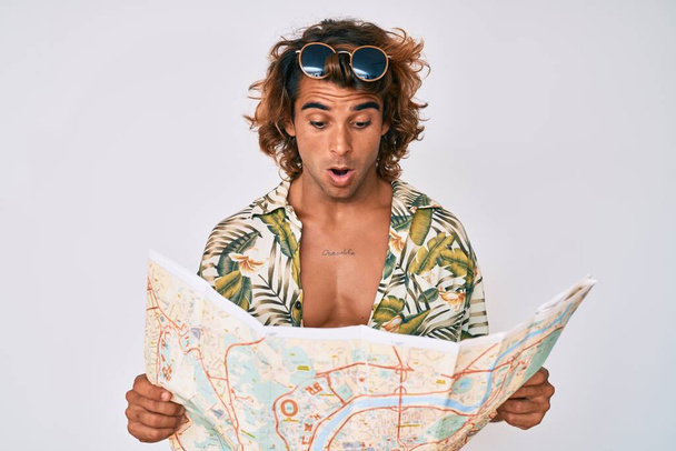 Joven hombre hispano con el estilo de verano sosteniendo el mapa en la cara de choque, mirando escéptico y sarcástico, sorprendido con la boca abierta  - Foto, Imagen