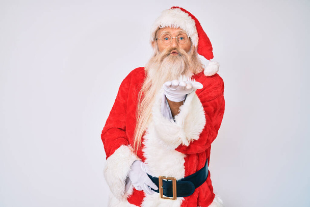 Ein alter älterer Mann mit grauen Haaren und langem Bart im Weihnachtsmann-Kostüm mit Hosenträgern, der in die Kamera blickt und einen Kuss mit der Hand in die Luft pustet, ist schön und sexy. Liebesausdruck.  - Foto, Bild