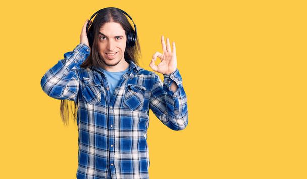 Homem adulto jovem com cabelos longos ouvindo música usando fones de ouvido fazendo ok sinal com os dedos, sorrindo amigável gesto excelente símbolo  - Foto, Imagem