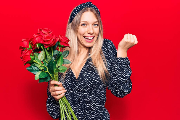 Young blonde woman holding flowers screaming proud, celebrating victory and success very excited with raised arm  - Fotoğraf, Görsel