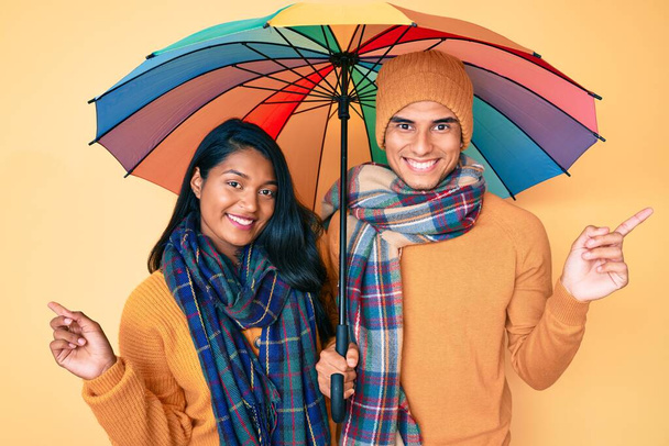 Schönes lateinisches junges Paar unter buntem Regenschirm lächelt glücklich und zeigt mit Hand und Finger zur Seite  - Foto, Bild