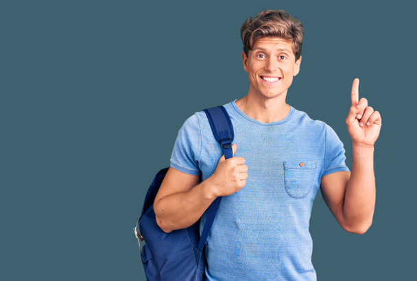 Jeune homme beau tenant un sac à dos étudiant surpris par une idée ou une question pointant du doigt avec un visage heureux, numéro un  - Photo, image