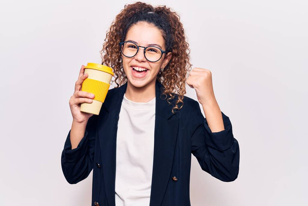 Beautiful kid girl with curly hair wearing business clothes and glasses holding takeaway cup of coffee screaming proud, celebrating victory and success very excited with raised arms  - Photo, Image